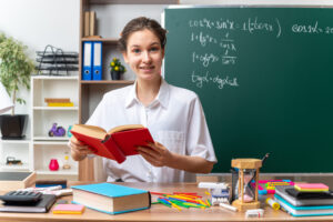 smiling-young-blonde-female-math-teacher-sitting-desk-with-school-tools-holding-book-classroom-scaled-1.jpg